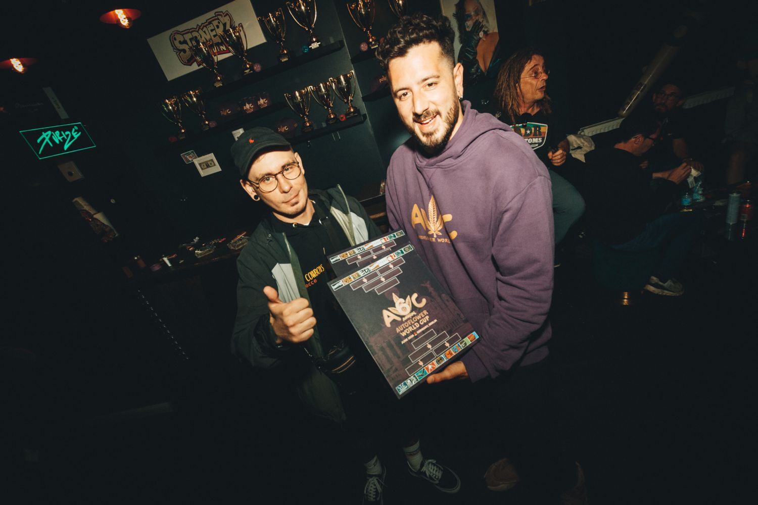 Timm Terpentino and a friend holding an Autoflower World Cup judging kit beside the trophy shelf