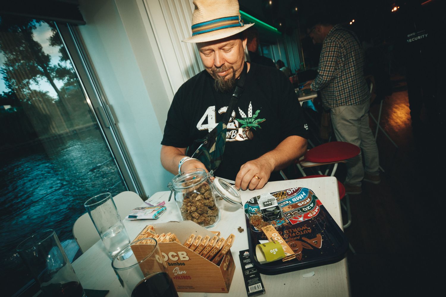 A man in a straw fedora and black cannabis-leaf t-shirt reaches into an open glass jar of cannabis buds at a white table set with a rolling tray, OCB bamboo papers and filters, and glasses of water, seated beside a window overlooking water at dusk.