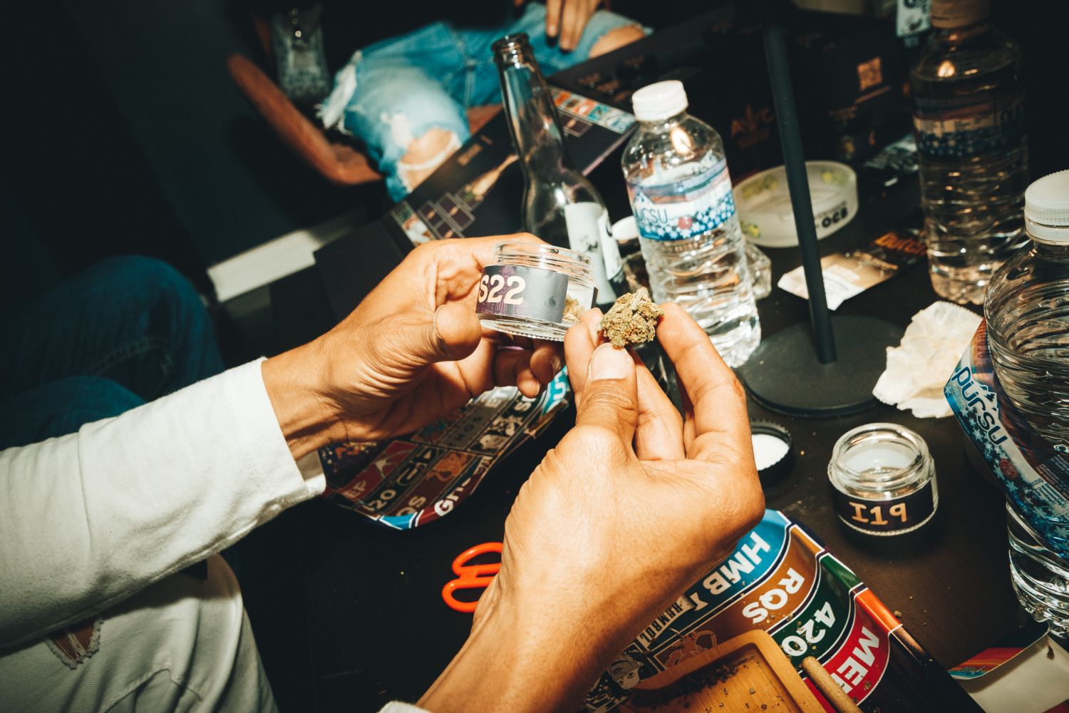 Examining a cannabis bud held between their fingers over a numbered Autoflower World Cup sample jar (S22), surrounded by water bottles, a rolling tray, and Humboldt Seeds packaging