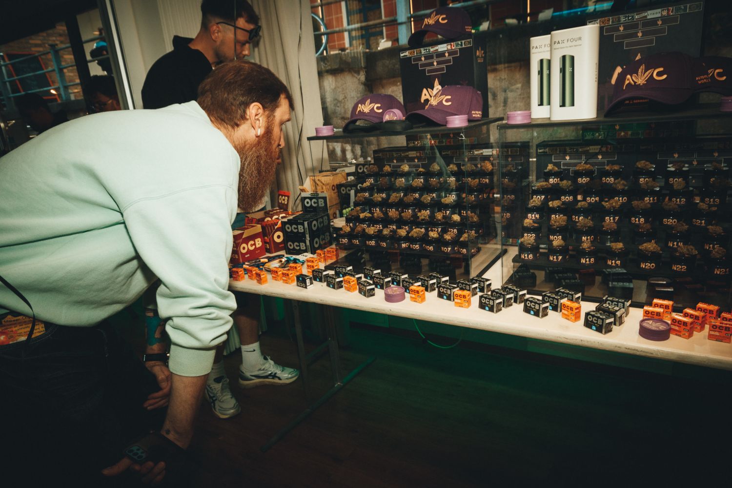 Bearded attendee leaning in to inspect rows of numbered cannabis entries beside OCB papers at the Autoflower World Cup