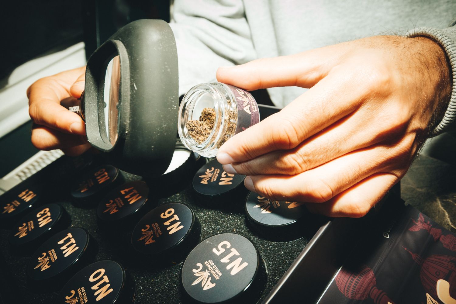 Judge examining a cannabis bud through a magnifying loupe at the Autoflower World Cup, with numbered black sample jars (N10&ndash;N18) on the table