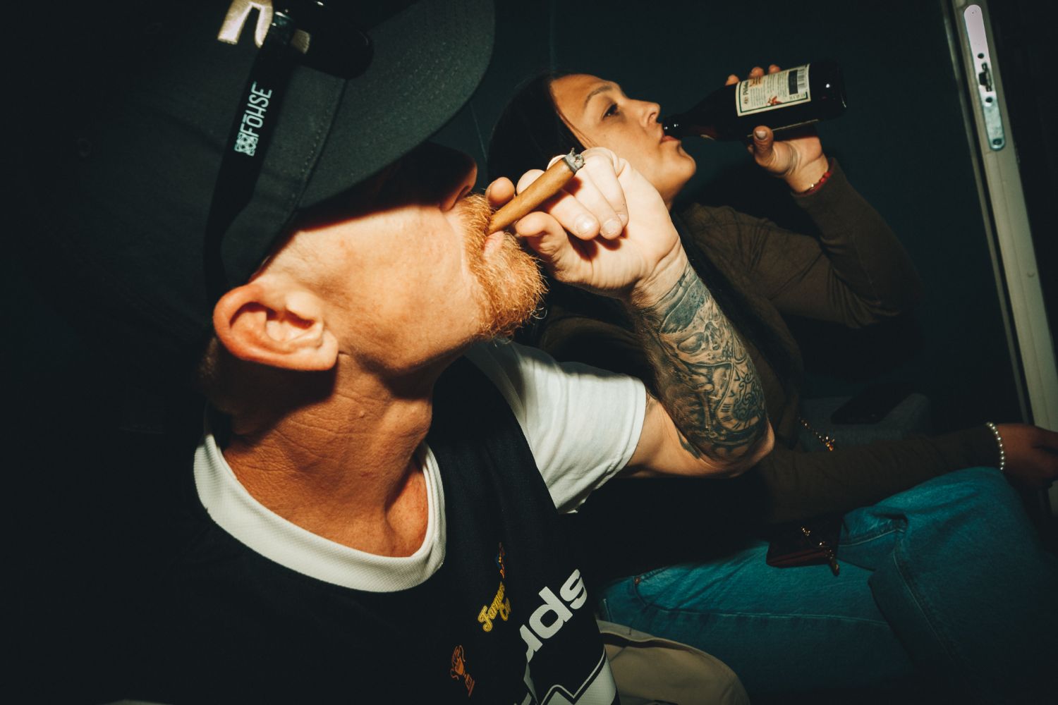 A bearded man in a black cap tilts his head back to light a cigar while a woman with a tattooed arm drinks from a beer bottle behind him in a darkly lit room.